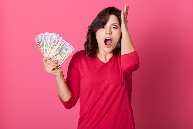 Amazed young woman holding bunch of money banknotes, celebrating profit, being shocked, keeps mouth widely opened and touching her head with palm, looks very surprised.