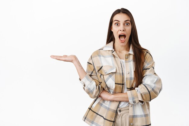 Amazed young woman holding something in open palm, display an awesome item, gasping excited and impressed with item, standing over white wall