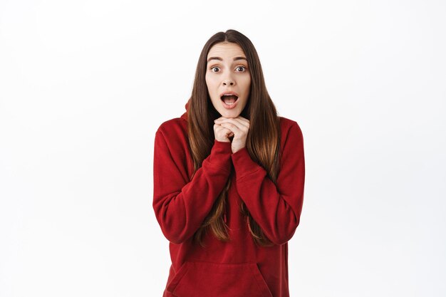 Amazed young woman listen to story with excitement, gasping and looking fascinated, watching something amazing and interesting, standing in red hoodie against white background