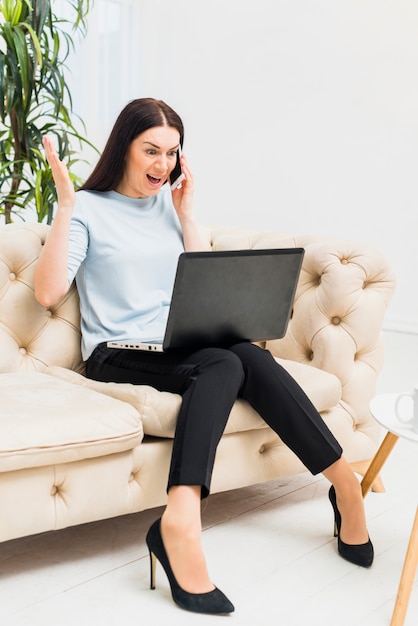 Amazed young woman sitting on couch with laptop and talking by phone 