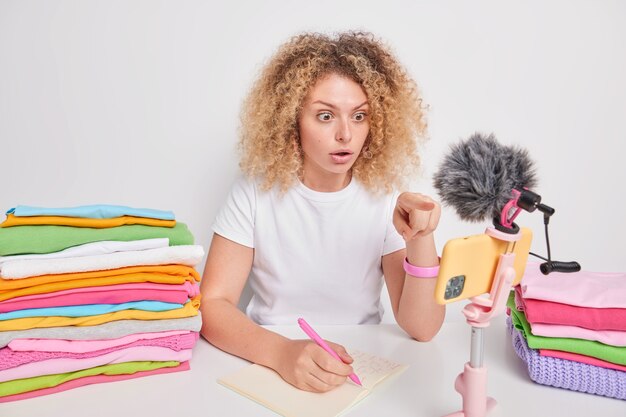 Amazed young woman with curly hair indicates at smartphone camera