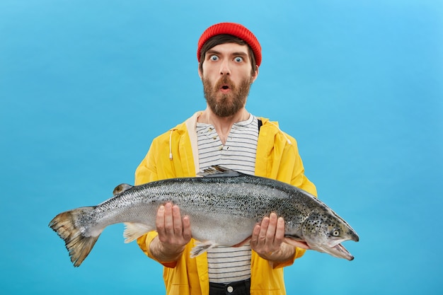 Amazement and unexpectedness concept. Shocked young fisherman with thick beard looking with bugged eyes and jaw dropped while holding huge fish not believing that he could catch it by himself