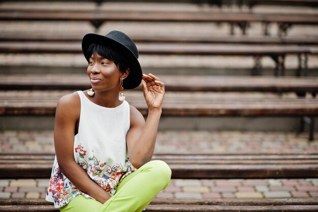 緑のズボンとベンチでポーズをとった黒い帽子の素晴らしいアフリカ系アメリカ人モデルの女性