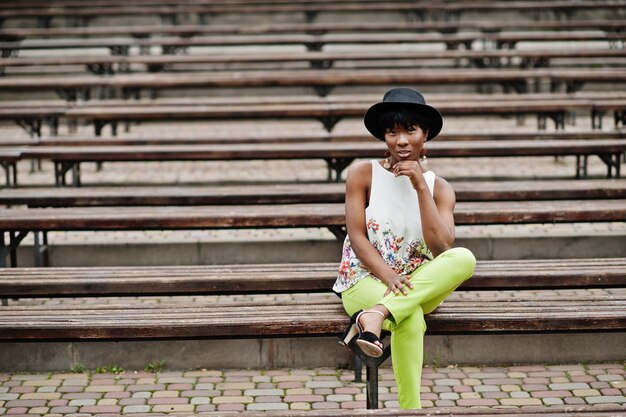벤치에 포즈를 취한 녹색 바지와 검은 모자를 쓴 놀라운 아프리카계 미국인 모델 여성