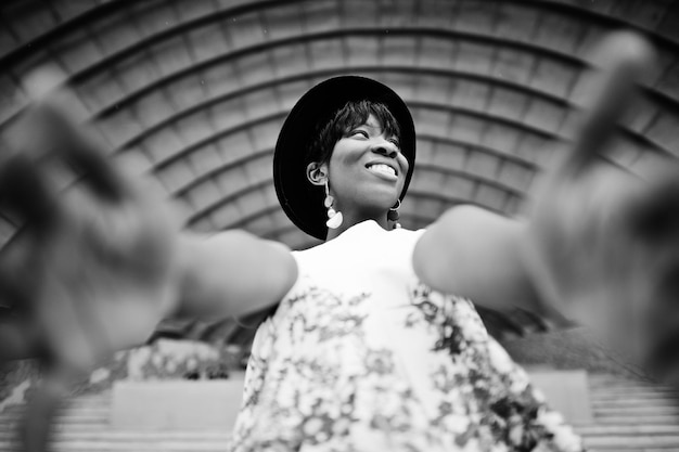 Incredibile donna modello afroamericano in pantaloni verdi e cappello nero in posa all'aperto contro la sala dell'arena che tiene la fotocamera