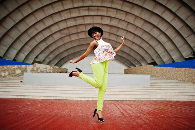 緑のズボンと黒い帽子の驚くべきアフリカ系アメリカ人モデルの女性がアリーナホールに対して屋外でポーズをとった空中ジャンプ