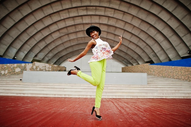 Amazing african american model woman in green pants and black hat posed outdoor against arena hall jump in the air