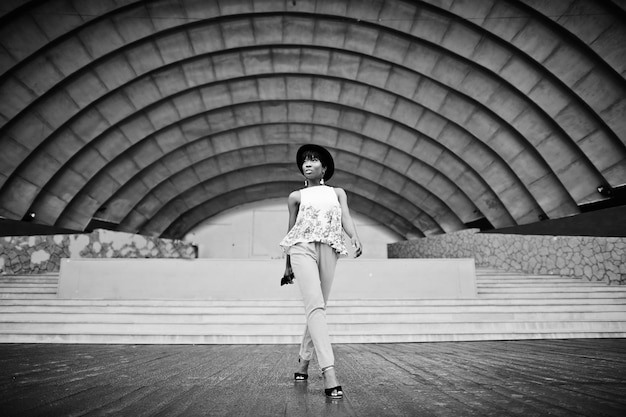Amazing african american model woman in green pants and black hat posed outdoor against arena hall