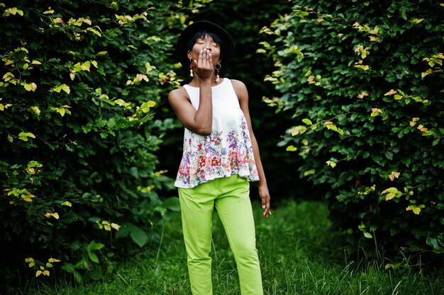 공원에서 다른 감정으로 포즈를 취한 녹색 바지와 검은 모자를 쓴 놀라운 아프리카계 미국인 모델 여성은 공기 키스를 보여줍니다
