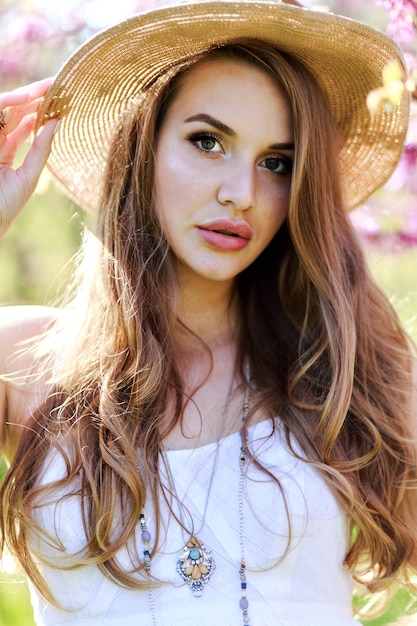 Incredibile attraente giovane donna con i capelli lunghi, in cappello, vestito di luce bianca godendo la giornata di sole primaverile in giardino su sfondo di fioritura sakura.