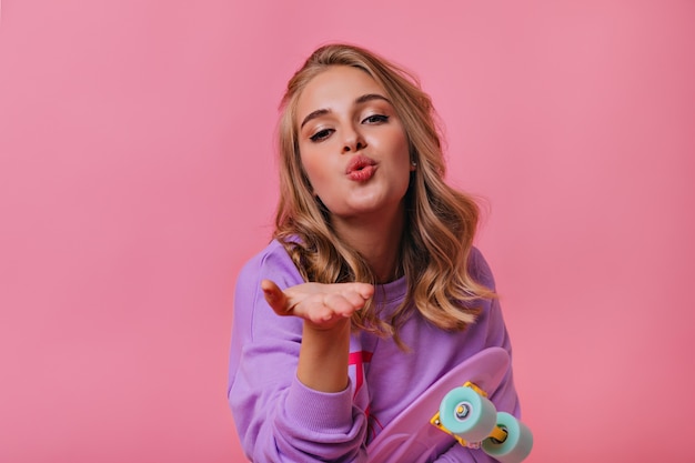 Incredibile ragazza bionda con trucco alla moda in posa con lo skateboard. adorabile signora caucasica che invia un bacio d'aria su pastello.