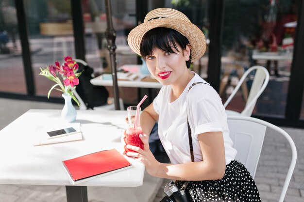 Amazing blue-eyed girl with dark hair under straw hat resting in cafe at the table with notebook, phone and flowers on it