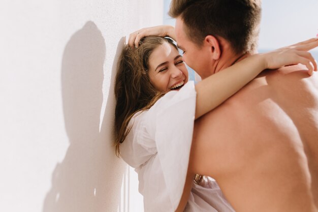 Amazing brunette girl in good mood embracing her boyfriend and smiling in sunny morning. Portrait of young woman and man hugging and playfully dancing in front of white wall.