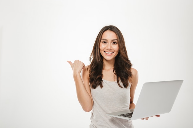 amazing businesswoman holding silver laptop and pointing finger sideways with smile, over white wall