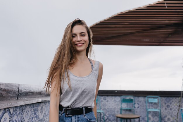 Удивительная кавказская девушка в повседневной одежде позирует с вдохновенной улыбкой. Фотография возбужденной женской модели, стоящей в кафе на крыше.