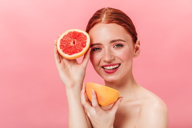 カットグレープフルーツを保持している驚くべき白人の女の子。ピンクの背景にポーズをとる柑橘類と素晴らしい裸の女性のスタジオショット。