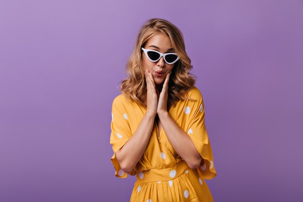 Amazing caucasian girl posing in trendy sunglasses. Indoor portrait of charming young woman in yellow attire isolated on purple.