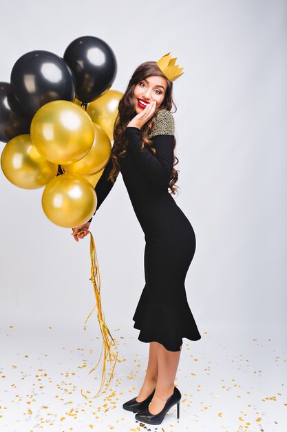 Удивительная жизнерадостная стильная женщина в черном вечернем роскошном платье и желтой короне на голове празднует новый год, улыбается и держит желтые и черные воздушные шары, красные губы, удивленное лицо.