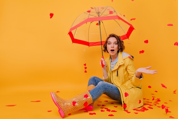 Удивительная кудрявая девушка с зонтиком, выражающая изумление во время сердечного дождя. Студия сняла милую брюнетку женскую модель в резиновых туфлях, позирующих в день святого валентина.