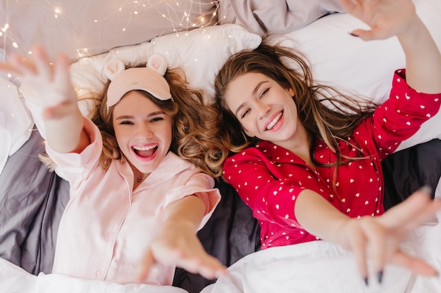 Amazing curly lady in trendy eyemask lying in bed with sister. Overhead portrait of two girls having fun before sleep.