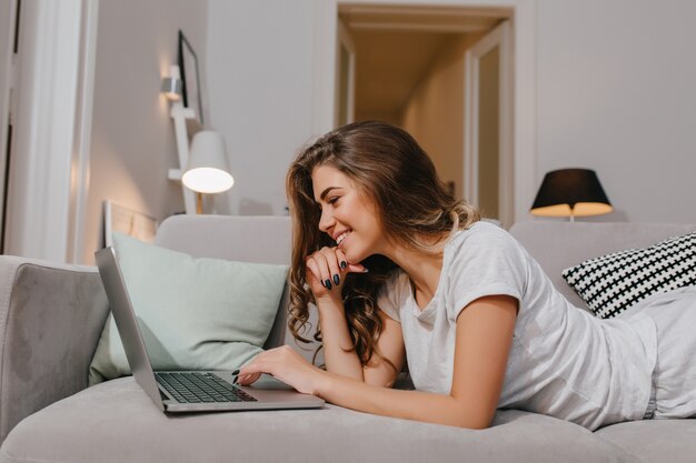 Amazing curly woman with black manicure lying on couch and typing on laptop keyboard