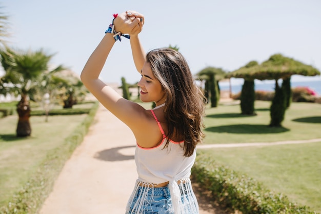Amazing cute girl with brown shiny hair gladly posing with hands up. Slim graceful young woman in white shirt smiling and dancing in park on summer resort in sunny morning