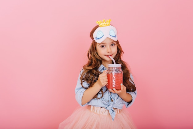 Amazing cute little girl with princess mask on head, long brunette hair drinking juice from glass and looking to camera isolated on pink background. Little happiness, expressing true positive emotions