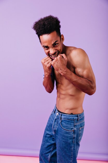 Amazing dark-haired african man posing without t-shirt. Indoor portrait of handsome curly boy isolated.