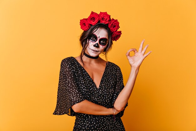 Amazing dead girl with scary makeup posing on orange background. Studio photo of lovely latin woman in halloween attire.