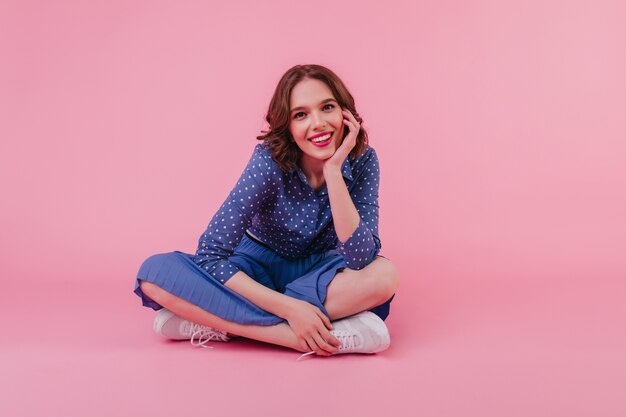 amazing european girl in trendy blouse sitting on pink wall. Beautiful white female model in skirt posing on the floor with legs folded.