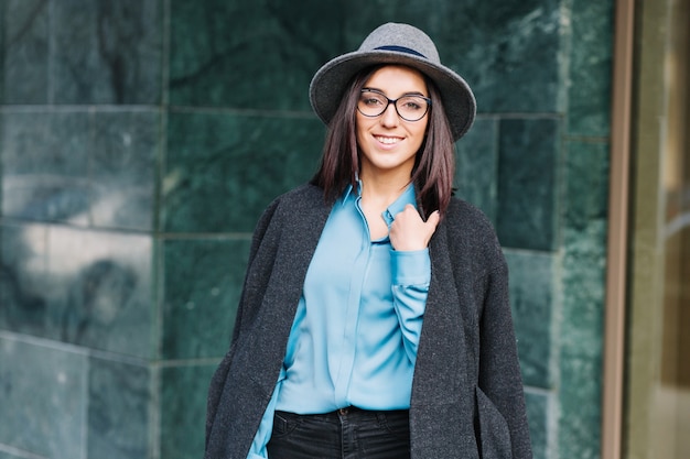 Amazing fashionable young woman in blue shirt, grey coat, hat walking outdoor on street in city. Brunette hair, black glasses, smiling, stylish businesswoman, elegant outlook.