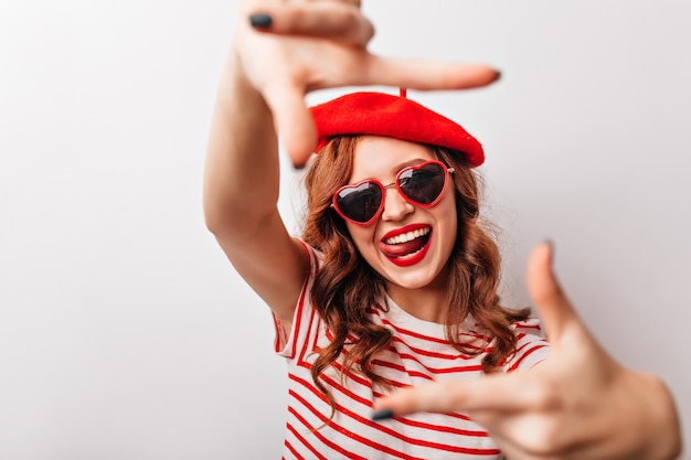 Amazing ginger girl in sunglasses having fun during photoshoot. Glad curly woman in french beret posing on white wall.