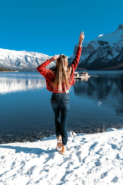 Amazing girl having fun outdoor by herself, enjoys the perfect nature view. Blue clear sky, big mountains and lake. Winter cold weather. Warm knitted red sweater and skinny jeans.