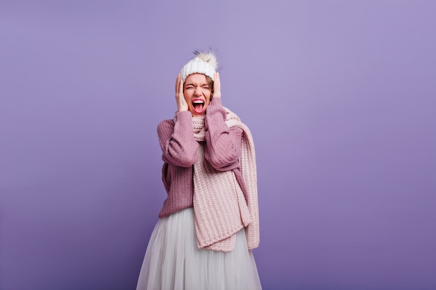 Free photo amazing girl in long knitted scarf screaming with eyes closed. magnificent european lady in stylish winter clothes posing on purple wall