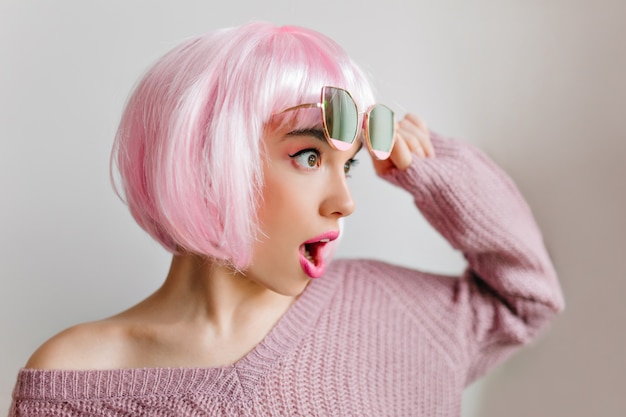 Удивительная девушка в розовом перуке позирует с удивлением и смотрит в сторону. Очаровательная женская модель в красочном парике, стоящем на светлой стене в очках.