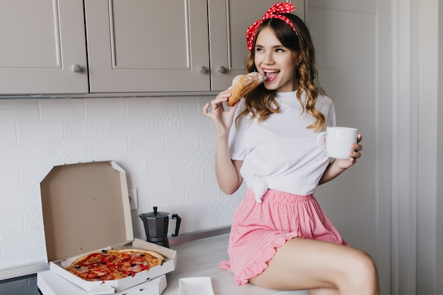 Amazing girl playfully eating croissant in morning. Lovely curly woman drinking coffee while sitting beside pizza.