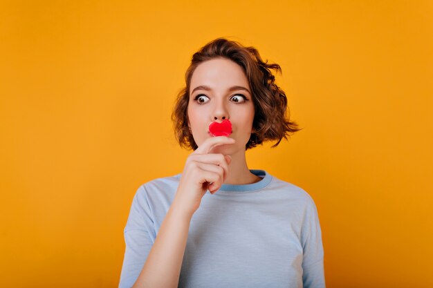 Amazing girl with big brown eyes posing with little red heart. Wonderful female model with trendy wavy hairstyle waiting for valentine's day.