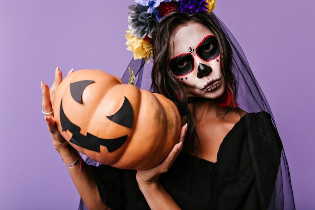 Free photo amazing girl with black bridal veil isolated on purple wall. indoor photo of evil lady in zombie attire holding halloween pumpkin.