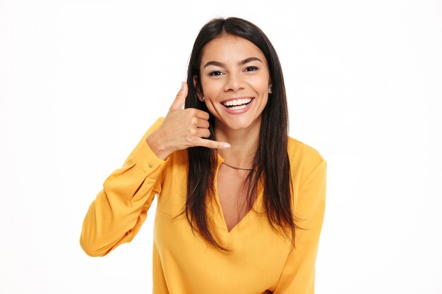 呼び出しジェスチャーを示す黄色のシャツで驚くほど幸せな若い女性。