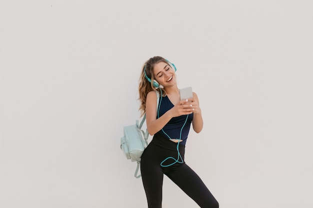 Amazing joyful pretty young woman in sportswear, with long curly hair chatting on phone. Workout, cheerful mood, happiness, listening to music through headphones, healthy lifestyle