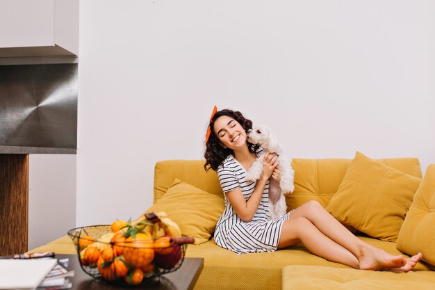 Amazing joyful young woman with cut brunette hair in dress chilling with dog on sofa in modern apartment. Having fun, home pets, joy, home comfort, cosy,  leisure, weekends