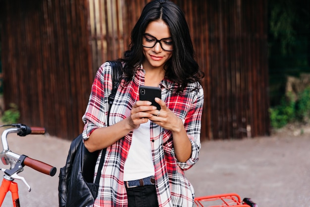 Удивительная латинская женская модель смотрит на экран телефона во время ожидания кого-то. Внешний портрет симпатичной женщины брюнетки, стоящей возле красного велосипеда.