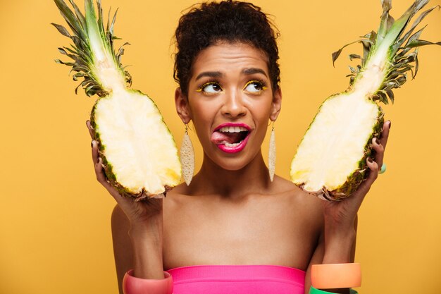 Amazing mulatto woman with colorful makeup looking upward and licking her lips while holding two parts of ripe appetizing pineapple isolated, over yellow