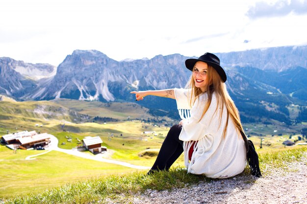 Amazing outdoor portrait of boho stylish woman posing at luxury resort with breathtaking mountains view, showing by her hand to Italian Dolomites.