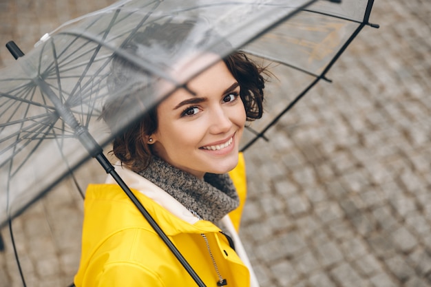 広い誠実な笑顔で透明な傘の下で黄色のコートに立っている若い女性の素晴らしい肖像画