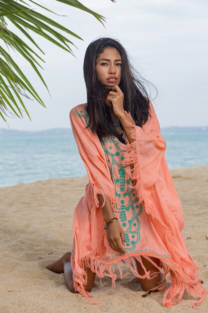 놀라운 섹시 황갈색 아시아 여자 팜 나무 아래 낙원 열 대 해변에서 포즈, 하얀 모래에 앉아 휴식과 휴일을 즐기는. 자수 장식의 보헤미안 드레스. 발리.
