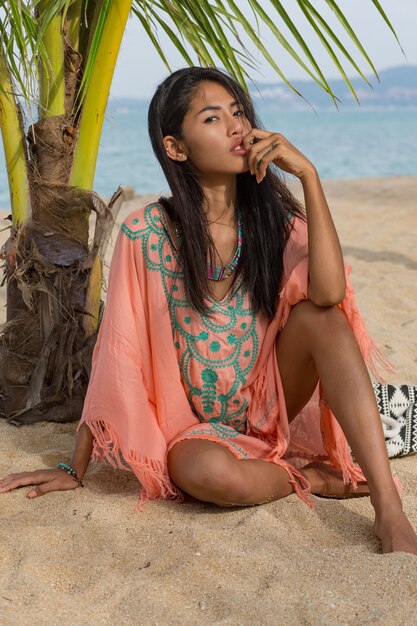 パムの木の下の楽園の熱帯のビーチでポーズをとって、白い砂の上に座って、リラックスして休日を楽しんでいる驚くべきセクシーな日焼けしたアジアの女性。刺繡の自由奔放に生きるドレス。バリ。