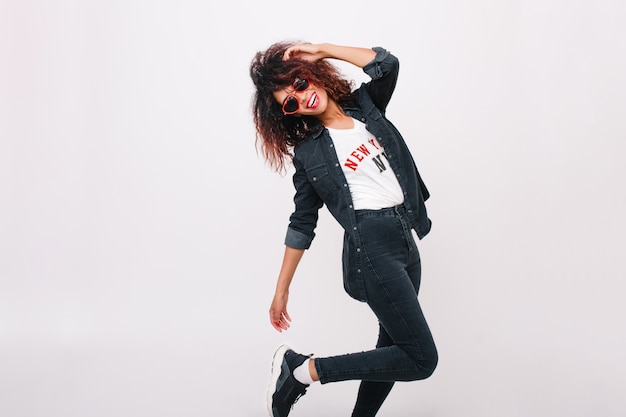 Foto gratuita incredibile modello femminile sottile in pantaloni neri in piedi su una gamba isolata con un sorriso. ragazza castana riccia che balla in scarpe da ginnastica alla moda e sorridente.