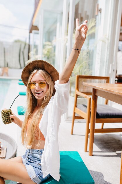 Amazing tanned woman with long hairstyle posing at resort with pineapple cocktail. lovely blonde woman in sunglasses and white shirt having fun in summer weekend.