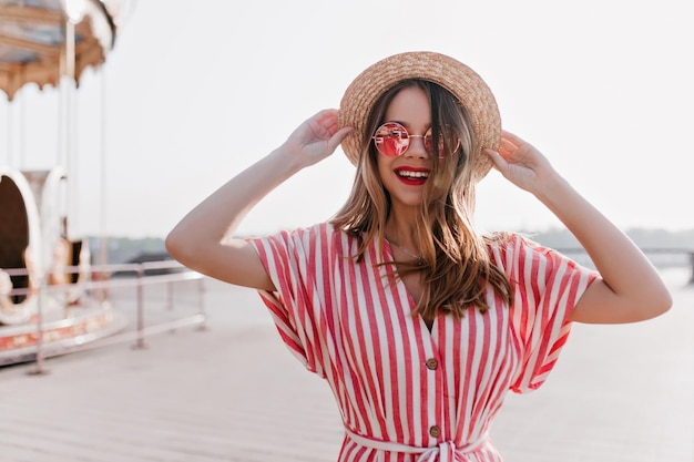 Foto gratuita incredibile modello femminile bianco che tocca il suo cappello sullo sfondo del cielo e sorridente elegante giovane donna in abito a righe godendo di un buon fine settimana estivo